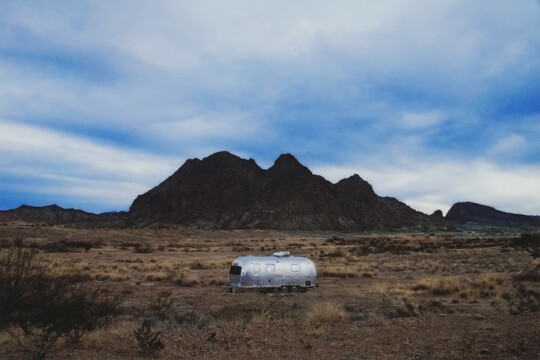 A color photograph of an Airstream-like trailer in a scrubby desert with mountains in the distance under a blue sky with soft white clouds.