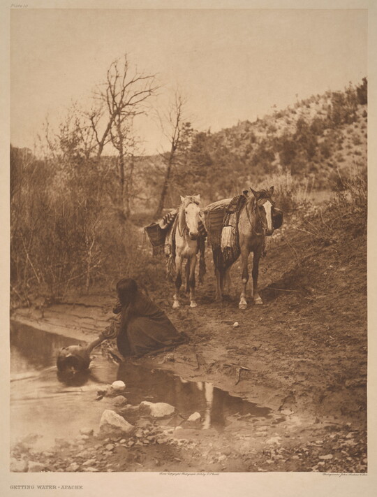 A sepia-toned photograph of an Indigenous person kneeling next to a stream to fill a jug with water as two packhorses wait in the background.
