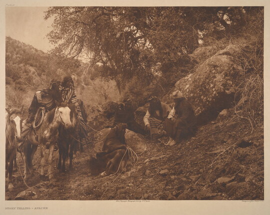 A sepia-toned photograph of a group of Indigenous people sitting on a grassy hill near their horses.
