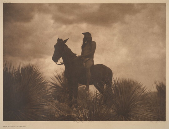 A sepia-toned photograph of an Indigenous person sitting atop a horse surrounded by yuccas; the low light makes the figures look almost like silhouettes.