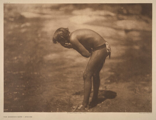 A sepia-toned photograph of a young Indigenous person leaning over a riverbank washing their face.