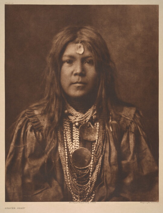 A sepia-toned portrait photograph of a young Indigenous person with long hair, a piece of jewelry on their forehead, multiple stings of beads around their neck, and wearing cultural dress.