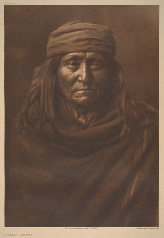 A sepia-toned portrait photograph of an older Indigenous person with long hair, wearing a cap, and wrapped in a solid-colored cloth.
