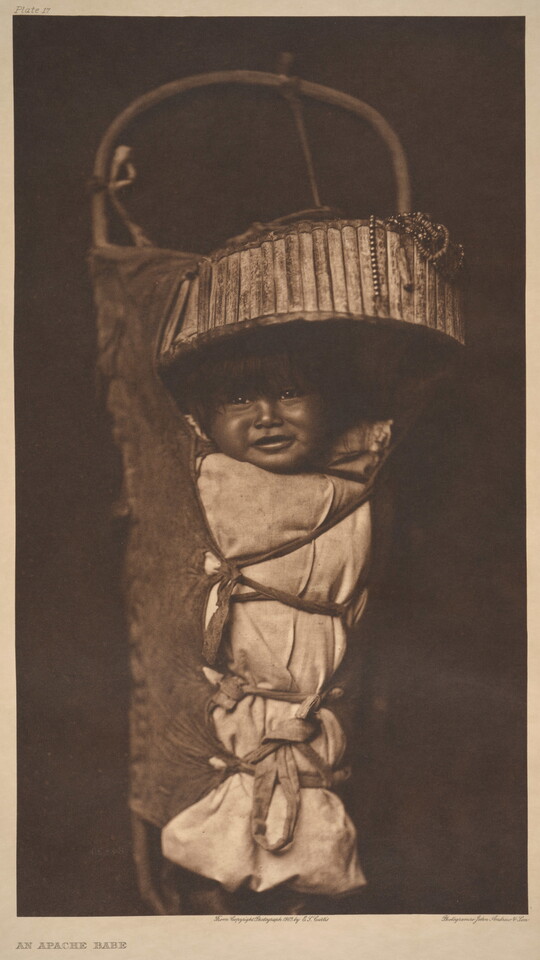 A sepia-toned photograph of a smiling Indigenous baby who is strapped into a traditional cradleboard.