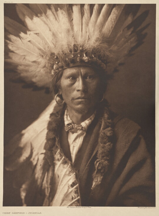 A sepia-toned photograph of a Native American man posed in a suit and feathered headdress.