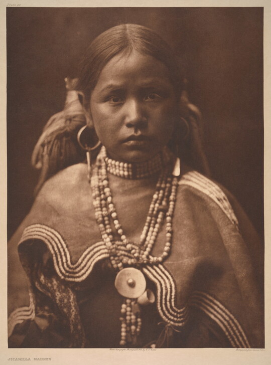 A sepia-toned portrait photograph of a young, Indigenous girl wearing a beaded necklace and cultural dress.