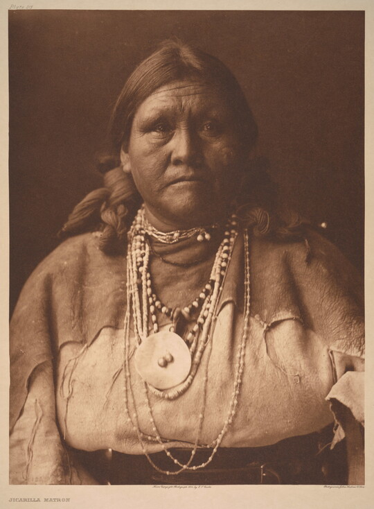 A sepia-toned portrait photograph of an older Indigenous woman with her hair bound on both sides of her head and wearing several beaded necklaces and traditional clothing.