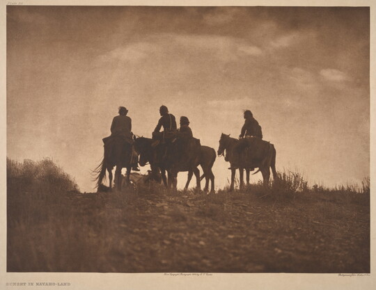 A sepia-toned photograph of the silhouettes of four people on horseback at the top of a hill.