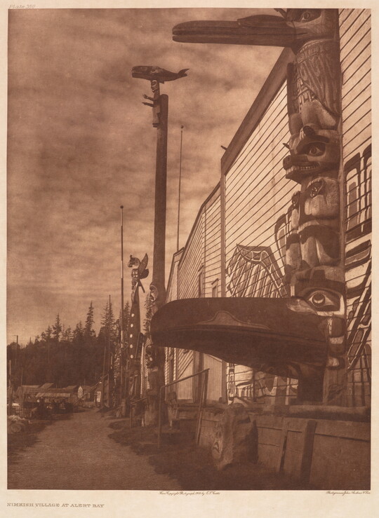 A sepia-toned photograph of several totem poles pressed next to a building with a mural painted on the side.