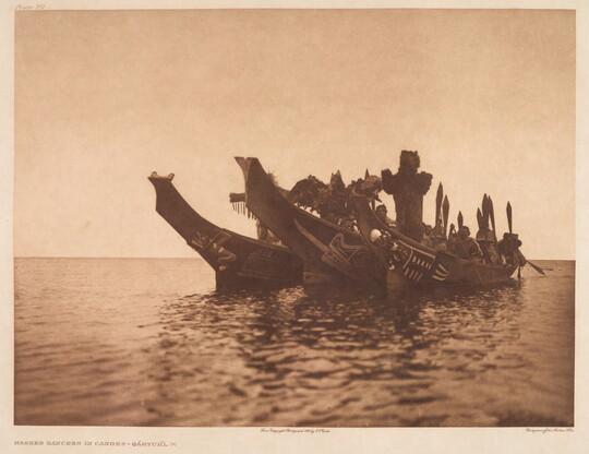 A sepia-toned photograph of three elaborately carved canoes side-by-side on a body of water with several masked Indigenous people in each canoe.