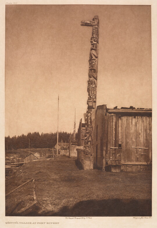 A sepia-toned photograph of a tall totem pole next to a wooden building with a forest in the background.
