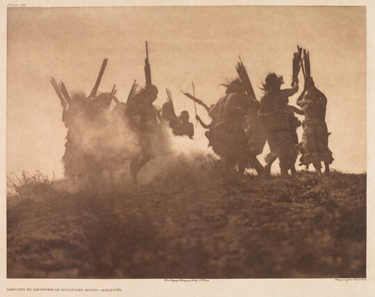A sepia-toned photograph of the silhouettes of a group of people stirring up dust as they dance in a circle.