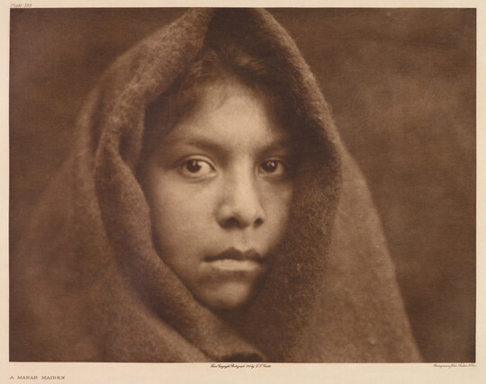 A sepia-toned portrait photograph of a young Indigenous girl wearing a cloth over her head, exposing only her face and hair above her forehead.