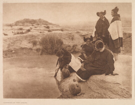 A sepia-toned photograph of a group of five Indigenous women wearing traditional dress sitting and standing on the edge of a spring.