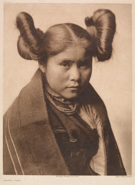 A sepia-toned portrait photograph of a young Indigenous woman with her hair split into two large buns on both sides of her head wearing traditional clothes and necklaces.