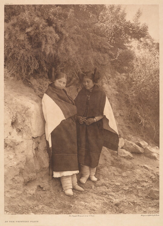 A sepia-toned photograph of two Indigenous women in traditional clothing leaning on a rock that has vegetation growing off the top of it.