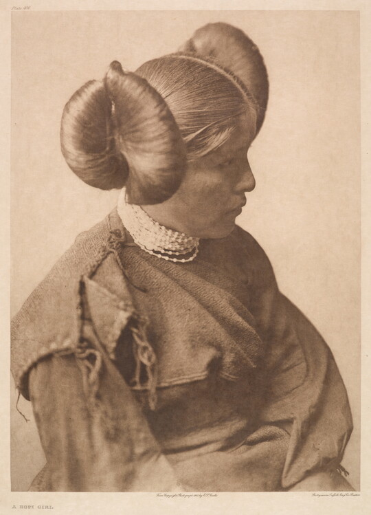 A sepia-toned portrait photograph of a young, Indigenous girl wearing a cultural hairstyle and a long sleeved dress.