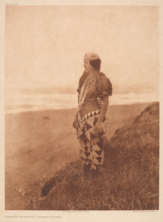 A sepia-toned photograph of a young Indigenous woman wearing traditional dress standing on a hill overlooking a plain.