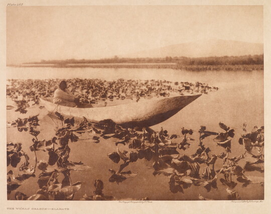 A sepia-toned photograph of an Indigenous person sitting in a canoe on water surrounded by vegetation.
