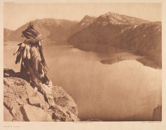 A sepia-toned photograph of an Indigenous man in traditional clothing and a feathered headdress sitting on a rock high above a lake at the base of mountains.