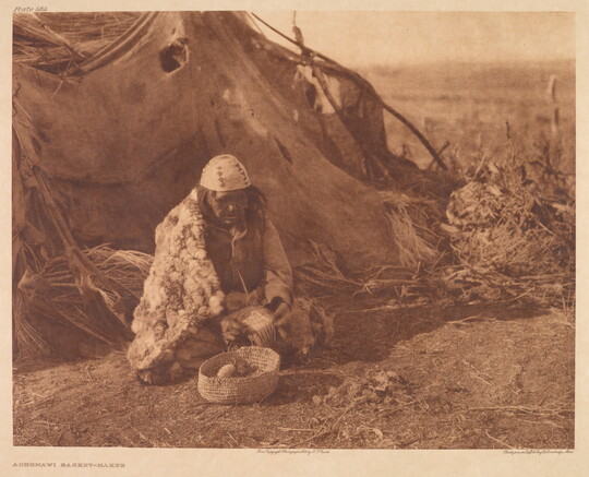 A sepia-toned photograph of an Indigenous person in traditional dress sitting on the ground outside of a hut with a woven basket in front of them.