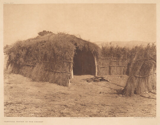 A sepia-toned photograph of a hut made of grass, brush, and wood on a dirt clearing.