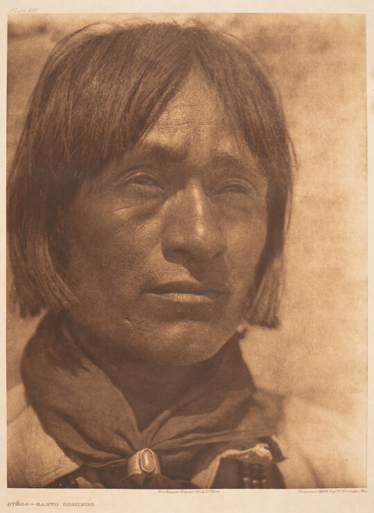 A close-up, sepia-toned portrait photograph of an Indigenous man with hair to his jawline, wearing a scarf around his neck held by a silver concho-style scarf slide.