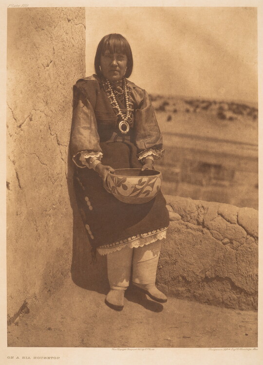 A sepia-toned photograph of an Indigenous woman in traditional dress holding a bowl seated on the ledge of a clay-style building.