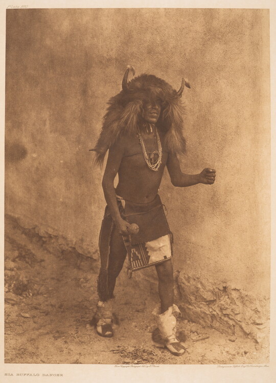 A sepia-toned photograph of an Indigenous man in cultural dress that includes a headpiece made of buffalo hide and horns.