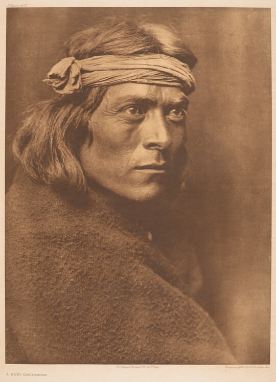 A sepia-toned portrait photograph of an Indigenous man wrapped in a blanket and wearing a cloth wrap on his head.