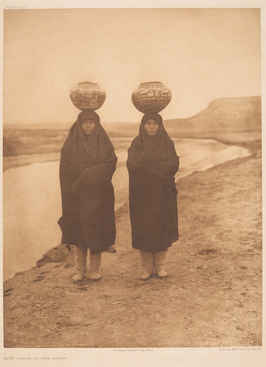 A sepia-toned photograph of two Indigenous women wrapped in blankets standing on a riverbed with water jugs balanced on their heads.