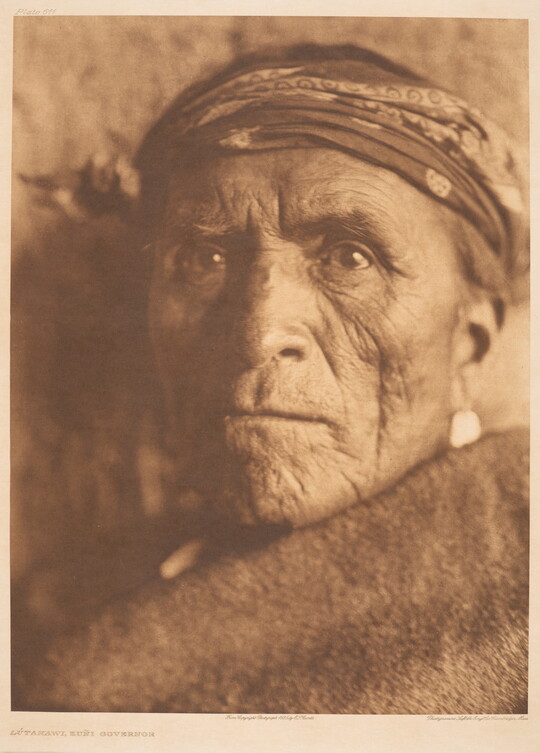 A sepia-toned portrait photograph of an older Indigenous person wearing a headband, and a solid-colored cloth wrapped around their shoulders and under their face.
