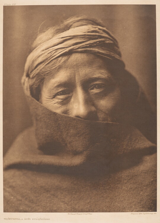 A sepia-toned portrait photograph of an older Indigenous person wearing headband; their face partially covered by what looks like a blanket.