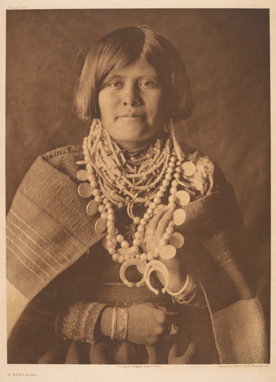 A sepia-toned portrait photograph of a young Indigenous person with short, bobbed hair, wearing many beaded necklaces, and a blanket wrapped around her shoulders.