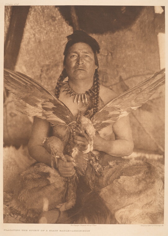 A sepia-toned photograph of a seated Native American man holding a large bird with its wings spread wide in one hand and a rattle in his other.