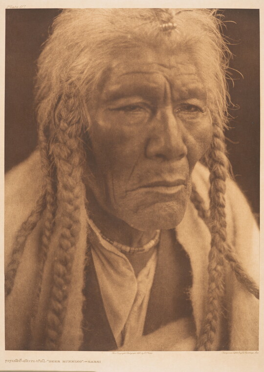 A sepia-toned portrait photograph of an elderly Native American man with long, gray hair in several braids gazing out.