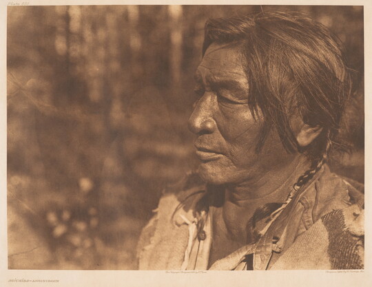 A sepia-toned photograph of a middle-aged Native American man's face in profile; short hair in two braids.