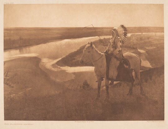 A sepia-toned photograph of a Native American man, in profile and wearing a full feathered headdress, on a horse looking off to a river in the background.