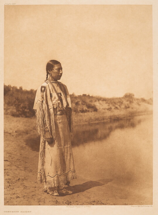 A sepia-toned photograph of an Indigenous woman with her hair pulled back into a braid, wearing fringed clothing that looks to be made of animal skins, standing on a shoreline.