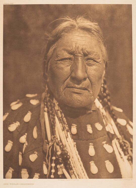 A sepia-toned portrait photograph of an older Indigenous woman wearing a braids, elk-tooth shirt, and multiple necklaces.