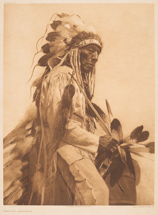 A sepia-toned portrait photograph of an older Indigenous man wearing a feathered headdress and cultural clothing.