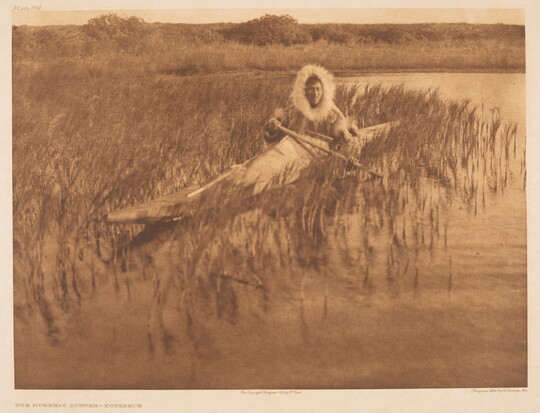 A sepia-toned photograph of an Indigenous person with a fur-lined hood pulled up around their head in a kayak among reeds on the water.