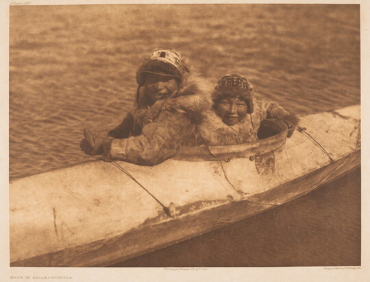 A sepia-toned photograph of two Indigenous people bundled up sitting in a kayak on the water.