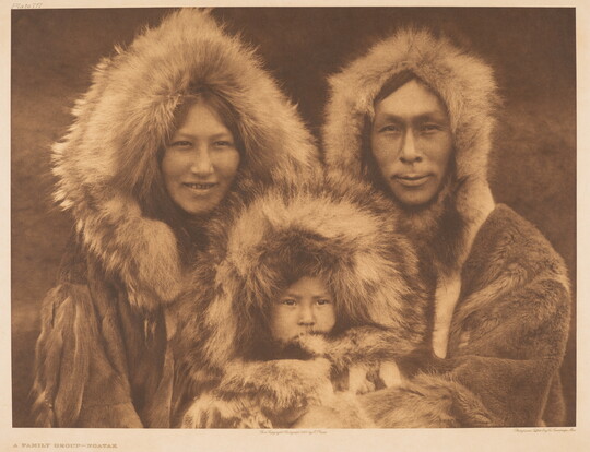A sepia-toned portrait photograph of an Indigenous family - woman, man, and baby - all wearing furs and looking at the camera.