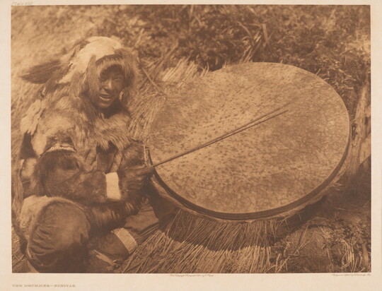 A sepia-toned photograph of an Indigenous person wearing furs squatting next to a large drum they are hitting with a stick.