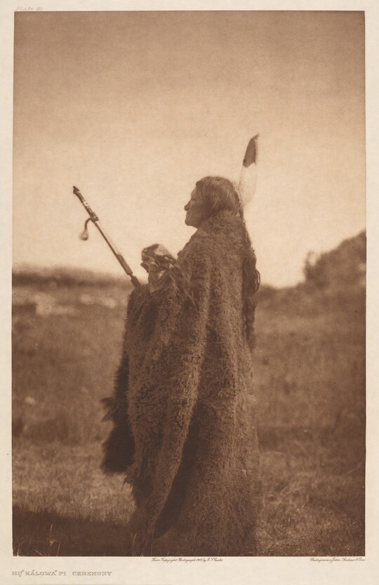 A sepia-toned photograph of an Indigenous adult standing on a grassy field wrapped in animal skins.