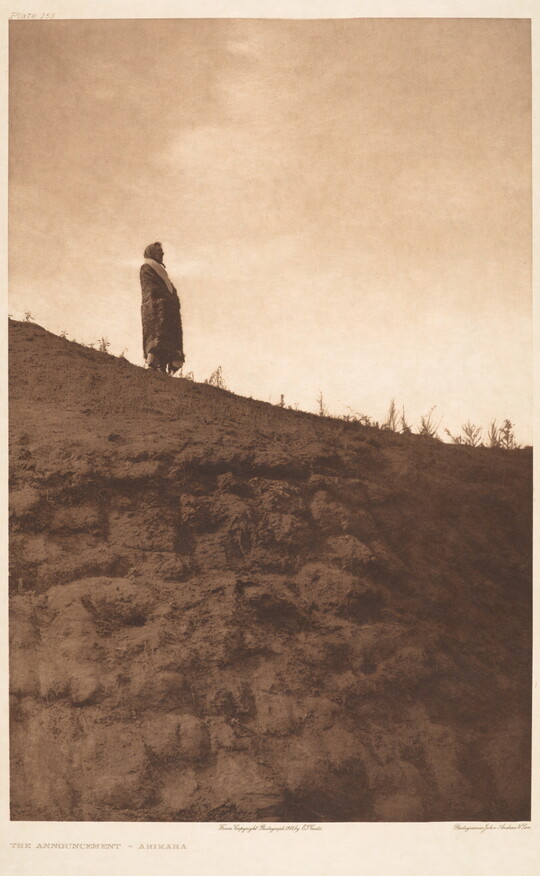 A sepia-toned photograph of an Indigenous person standing high on a hill with a blanket around their shoulders.