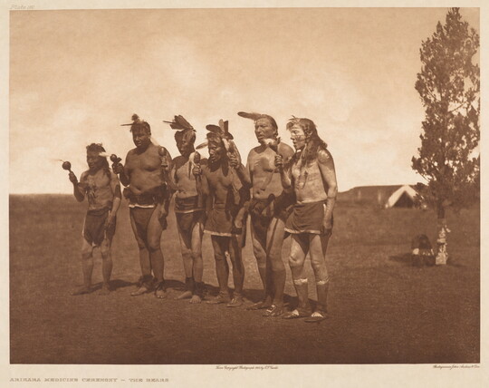 A sepia-toned photograph of six Native American men standing shoulder-to-shoulder, all holding something in their right hands.