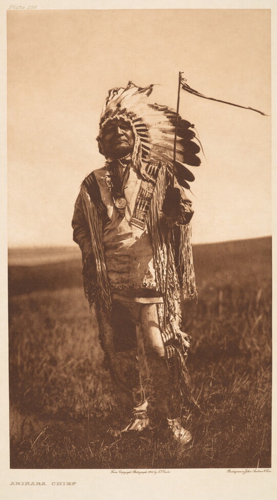 A sepia-toned photograph of an Indigenous man standing in a grassy field wearing a feathered headdress and ornately decorated traditional clothing.