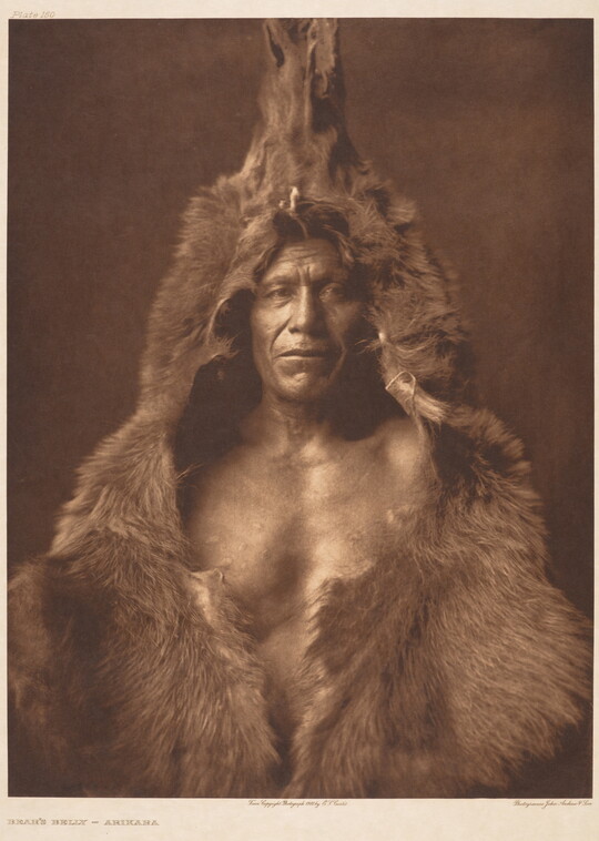 A sepia-toned portrait photograph of a Native American man wearing a bear skin over his head and shoulders.